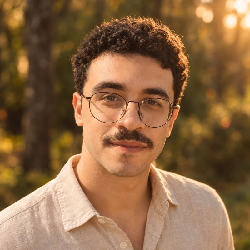 Portrait of Samuel Dutra: a smiling man with short curly dark hair, round glasses and a mustache, in a beige linen shirt, outdoors in a forest at golden hour.
